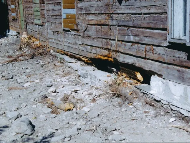 The image shows an old wooden house with a window and a door, surrounded by stones on the ground....