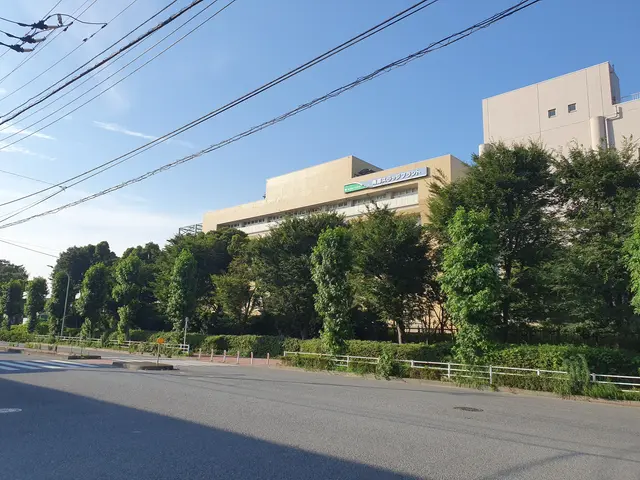 The image shows a road with a railing, plants, trees, light poles, and a building with a sign on...