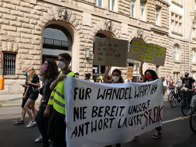 The image shows a group of people walking down a street, some of them holding placards and banners,...