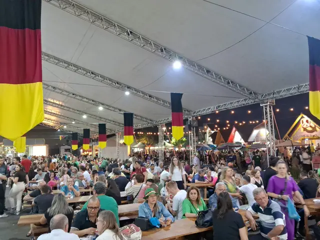 The image shows a large group of people sitting at tables under a tent at the Oktoberfest in...