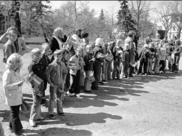 The image shows a group of people standing in a line on a dirt road, holding flags in their hands....