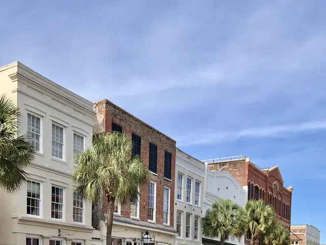 The image shows a city street with cars parked on the side of it, buildings with windows, trees,...