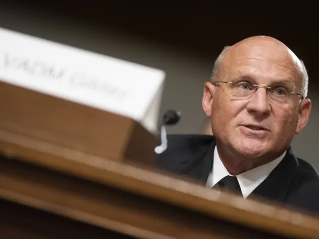 The image shows a man in a suit and tie sitting at a table with a name board and microphone in...