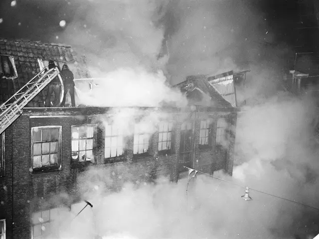 The image shows a black and white photo of a building engulfed in flames, with smoke billowing out...