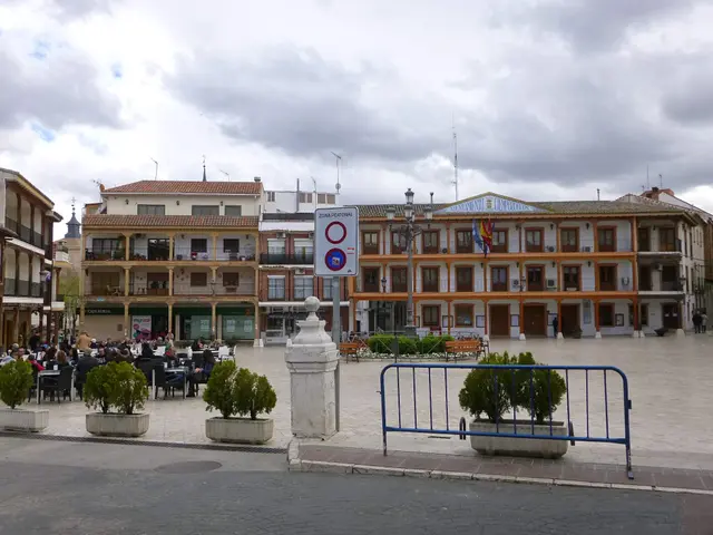 The image shows a bustling plaza mayor in the center of a city, with a group of people sitting on...