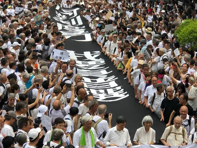 The image shows a large group of people walking down a street, holding banners and placards in...