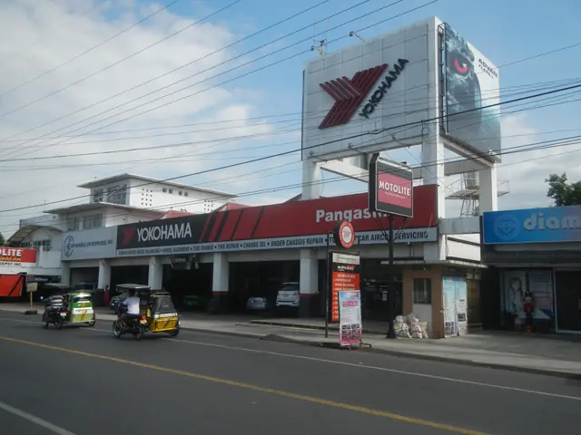 The image shows a bustling city street with vehicles driving on the road, buildings with name...
