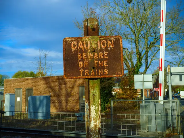 The image shows a caution sign on the side of a train track, surrounded by a metal fence, a group...