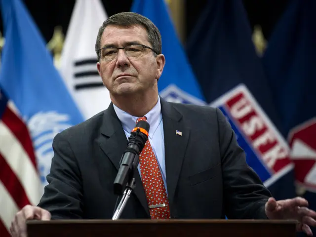 The image shows a man in a suit and tie standing at a podium with a microphone in front of him and...