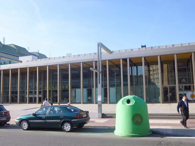 The image shows a green trash can sitting on the side of a street next to a building with glass...