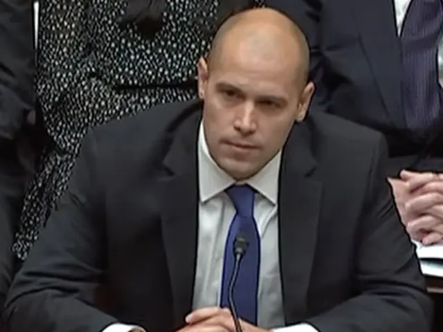 The image shows a man in a suit and tie sitting at a table with a name board, a bottle, a pen, and...