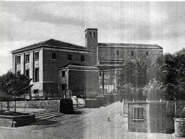 The image shows a black and white photo of a large building with windows, trees, steps, a fence,...