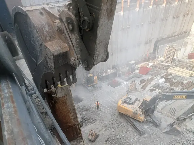 The image shows a construction site with a large excavator in the foreground and a building in the...