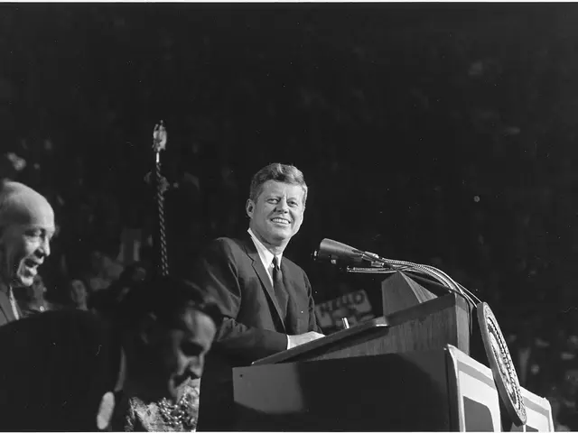 The image shows President John F. Kennedy standing at a podium in front of a crowd, wearing a...