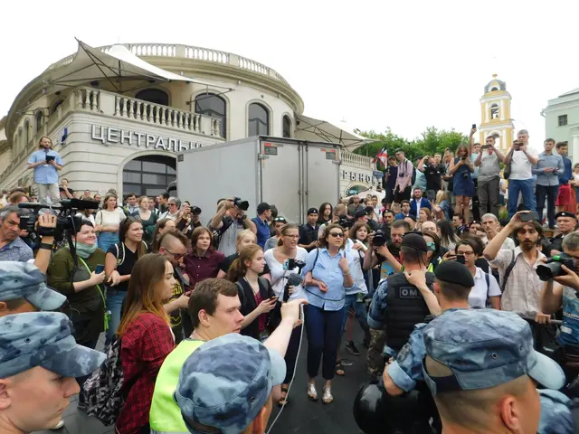 The image shows a large group of people standing in front of a building, some of them holding...