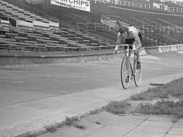 The image shows a man riding a bicycle down a street next to a stadium. He is wearing a helmet and...