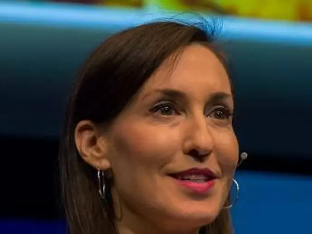 The image shows a woman in a white jacket and black top sitting down in front of a blue and yellow...