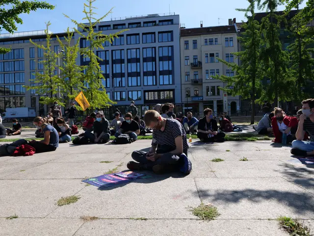 The image shows a group of people sitting on the ground in front of a building, surrounded by trees...