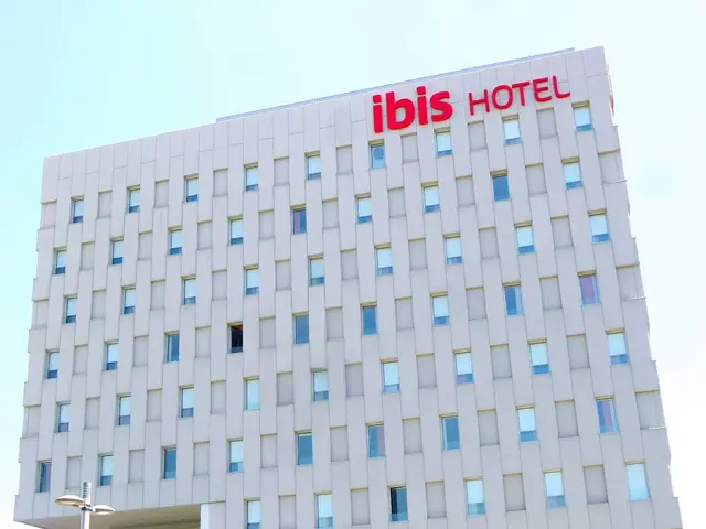 The image shows the ibis hotel in Berlin, Germany. It is a large building with windows, a street...