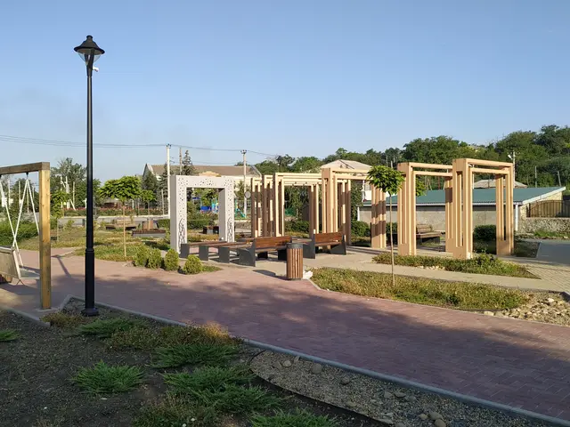 The image shows a park with a walkway lined with wooden poles, benches, plants, grass, stones,...