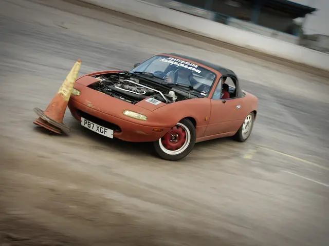 The image shows a person driving a red car on a race track, with a traffic cone in front of them....