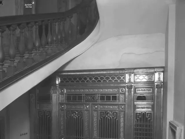 The image shows a black and white photo of a stairwell with a railing on the left side and a wall...