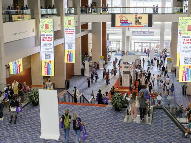 The image shows a large group of people walking through a large building with banners, houseplants,...