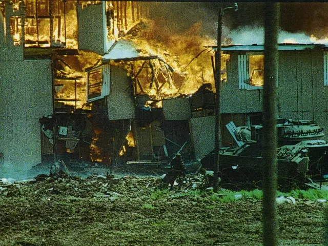 The image shows a house engulfed in flames with smoke billowing out of it, surrounded by poles,...