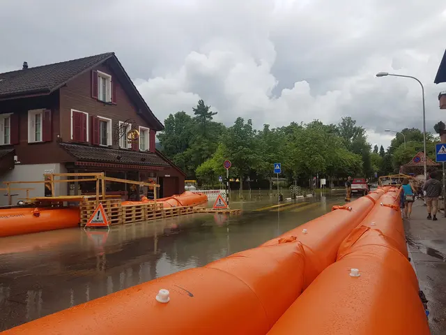 The image shows a flooded street with orange pipes in the middle of it. On the right side of the...
