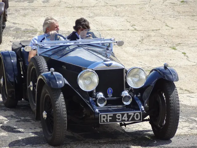 The image shows a man driving a vintage car down a street next to another car. The man is wearing a...