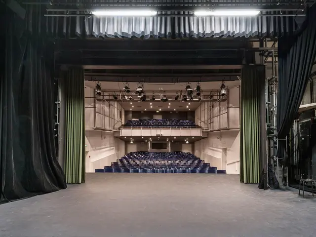 The image shows a large auditorium with black curtains and a stage, illuminated by lights on the...