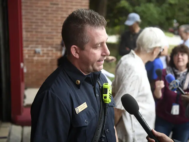 The image shows a man in a police uniform talking to a group of people, some of whom are holding...