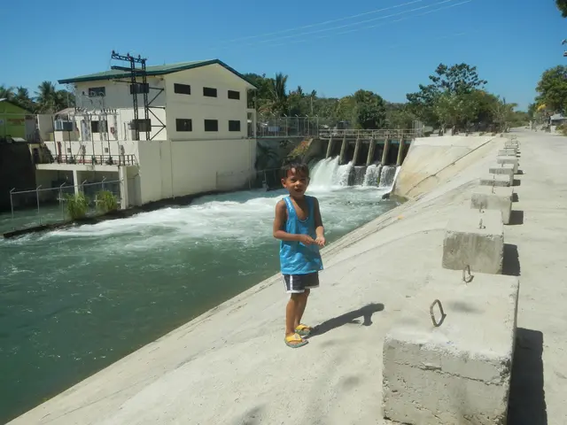 The image shows a young boy standing on top of a concrete wall next to a river, with a dam in the...