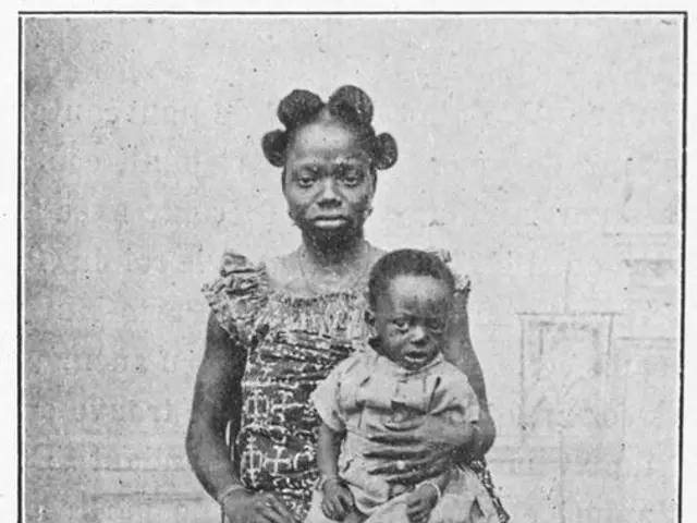 The image shows an old black and white photo of a woman sitting on a chair, cradling a baby in her...