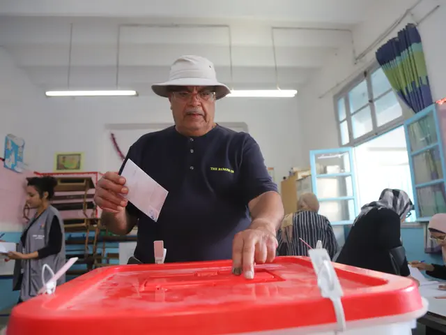 The image shows a man in a hat casting his vote at a polling station. He is standing in front of a...