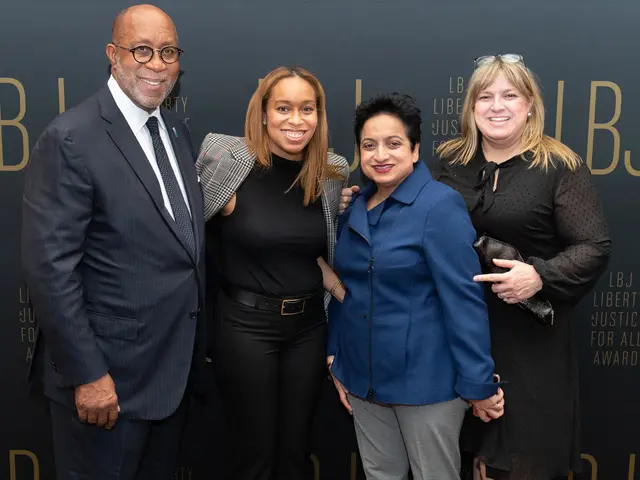 The image shows a group of four people standing next to each other on a red carpet, smiling and...