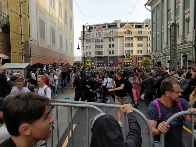 The image shows a large crowd of people standing in front of a metal barricade on a road surrounded...