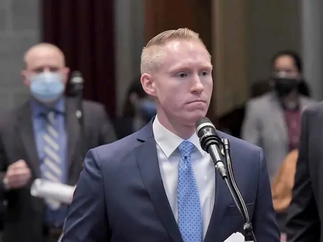 The image shows a man in a suit and tie standing at a podium, holding papers in his hands and...