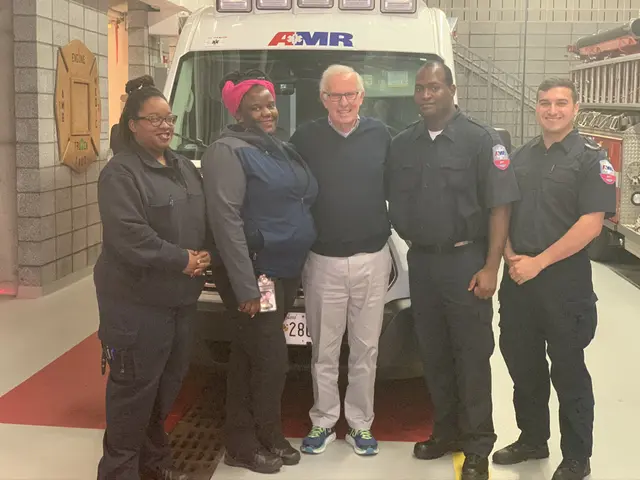 The image shows a group of people standing in front of an ambulance. The ambulance is parked on the...