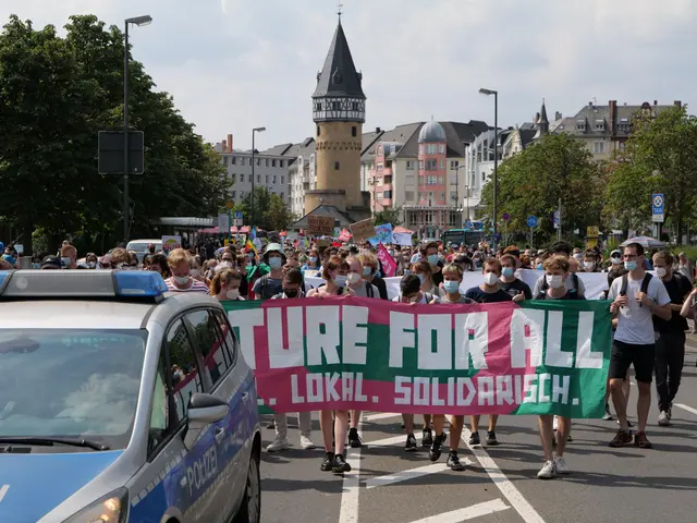 The image shows a large group of people walking down a street, holding a banner with the words...