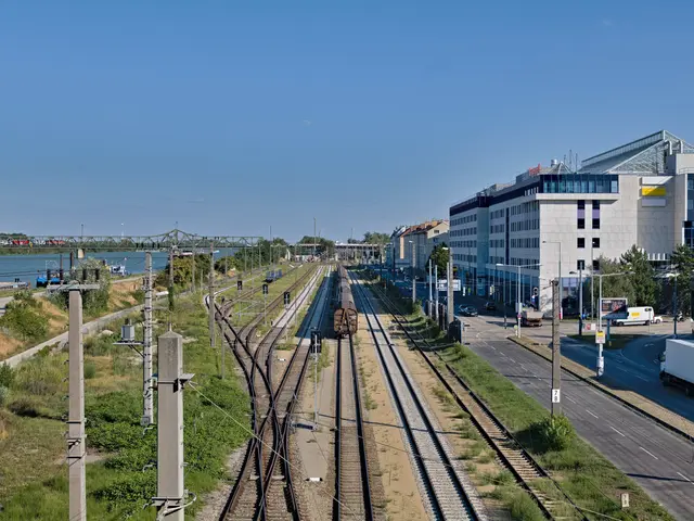 The image shows a train traveling down train tracks next to a city, with buildings, street poles,...