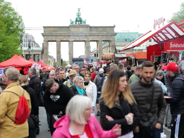 The image shows a crowd of people walking down a street in front of the Brandenburg Gate in Berlin,...