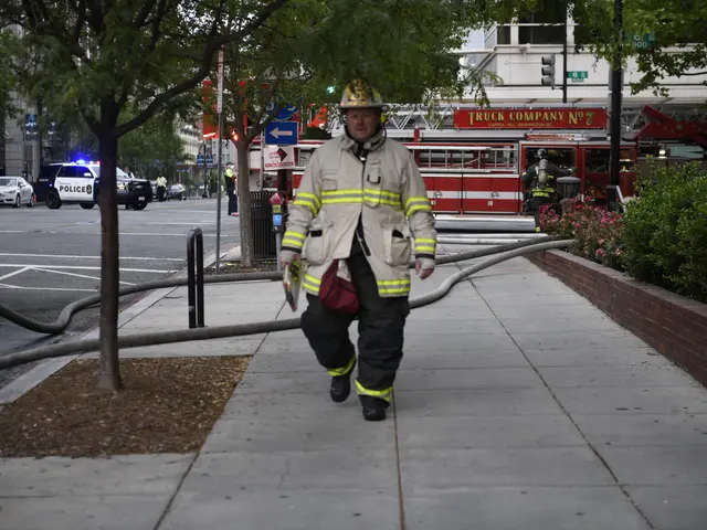 The image shows a firefighter walking down a sidewalk with a hose in his hand. He is wearing a...