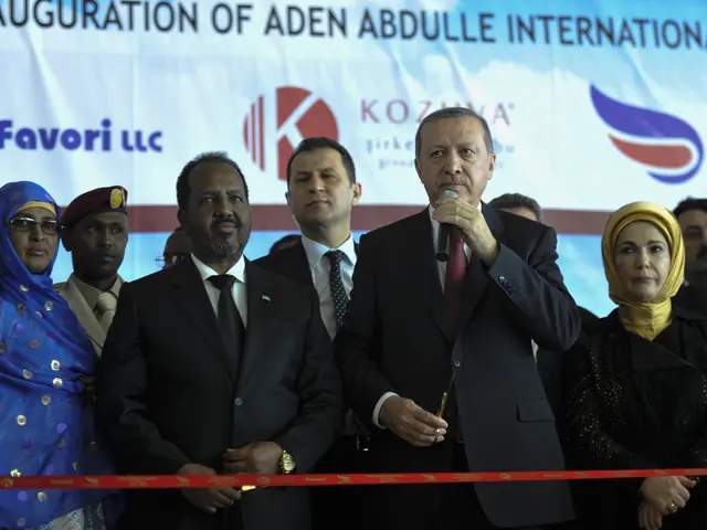 The image shows a group of people standing next to each other in front of a red ribbon, with a man...