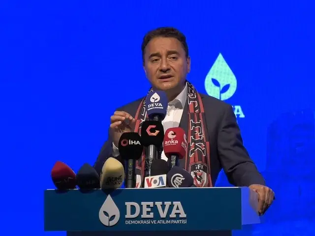 The image shows a man standing at a podium with microphones in front of him, wearing a coat and a...