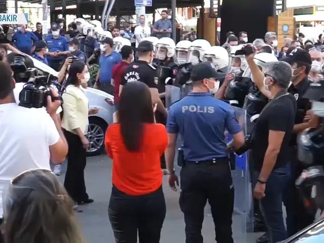 The image shows a group of people standing in front of a crowd of police officers wearing helmets...