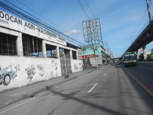 The image shows a city street with cars and trucks driving down it, a bridge on the right side,...