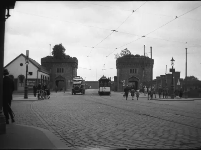 The image shows a black and white photo of a city street with a few vehicles and people on the...