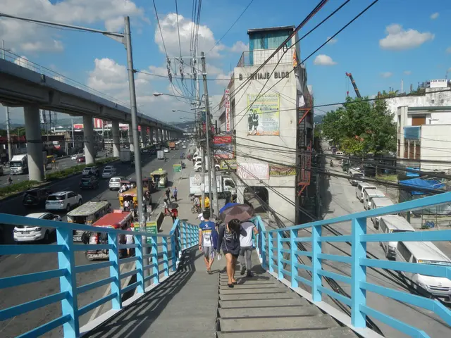 The image shows a group of people walking across a pedestrian bridge over a busy street. The bridge...