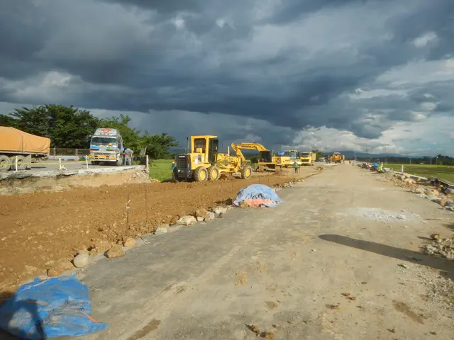 The image shows a dirt road with construction equipment on the side of it, including cranes,...